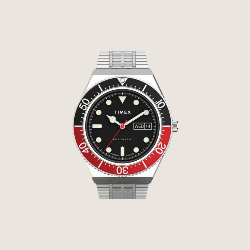 Timex M79 automatic with black dial, black and red dive bezel, day-date and stainless steel mesh bracelet, on a plain bone background